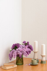 Fototapeta premium Vase with beautiful lilac flowers, candles, cup and books on table near light wall in room, closeup