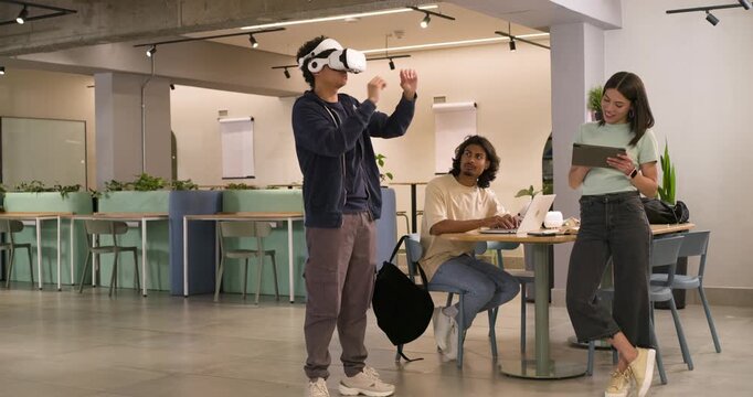 Diverse team watching male tester adjusting white VR headset demoing gestures with tablet in office
