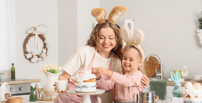 Happy mother and her cute daughter in bunny ears decorating Easter cake at home