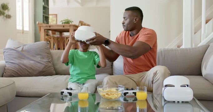 African American dad on sofa lifting VR headset adjusting on son while reaching controllers playing