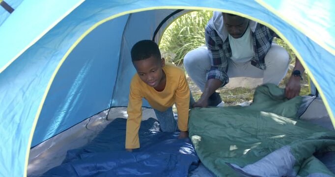 African-American father-son unfolding green bag and smoothing blue pad in blue tent preparing rest