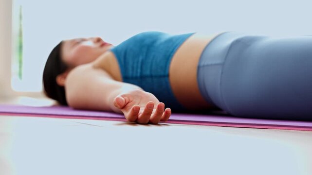 Relaxed young Asian woman lying back flat floor practicing yoga corpse pose savasana purple mat stretching bright studio calm peaceful resting expression feeling tranquil healthy fitness concept