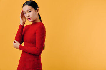 Obraz premium Young woman in red sweater with long sleeves poses thoughtfully against orange background. Model with long dark hair looks down with hand near head in studio shot.