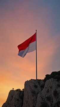 Indonesia or Monaco flag flies on a high pole on the edge of high rock hill against sunset sky
