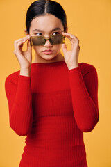 Naklejka premium Young woman in red sweater adjusting sunglasses on face with careful expression, posing against yellow background in studio for fashion portrait or lifestyle shoot.