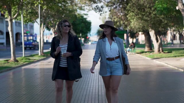 Two young stylish women walking towards the camera on a tree-lined city boulevard and talking in La Serena 