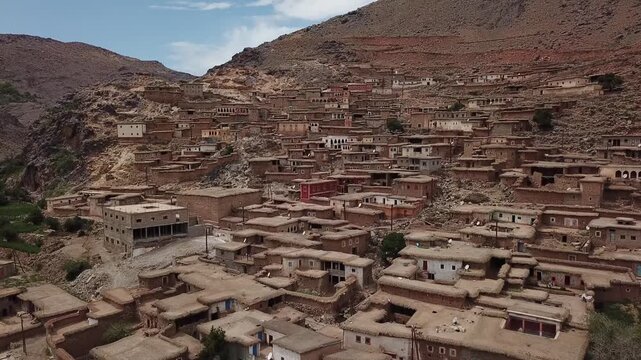 Aerial drone shot of a sprawling Berber village in the High Atlas. Highlights include dense indigenous earth architecture integrated into rugged rock strata and weathered mountain slopes.