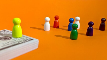 A bright yellow game piece stands prominently on a stack of currency, contrasting with a diverse group of colored pawns on an orange surface to illustrate concepts of wealth and social standing. © FaizAhmad