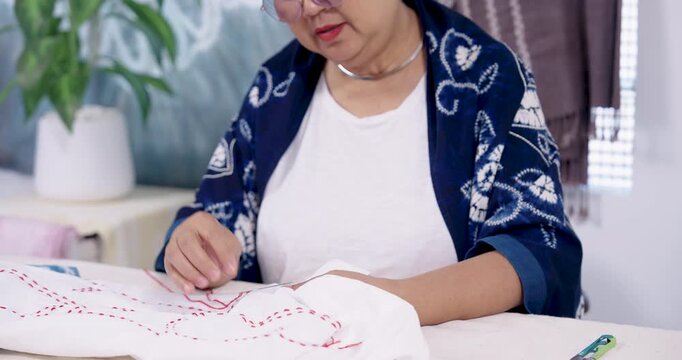 Senior Asian artisan leans forward, checks marked lines, then pierces cloth with needle to continue resist stitching for shibori tie dye, documenting slow living craft workflow for marketplace.