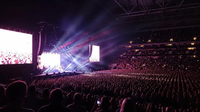 Concert crowd in a large stadium with stage and screens.