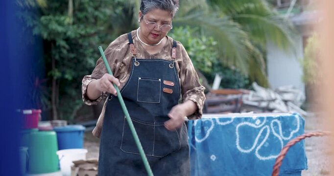Senior Asian woman lifts wet fabric bundle above basin, dye drips back while steam rises, revealing resist pattern after indigo dyeing, outdoor handmade textile workflow for small business sale.