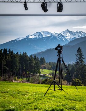 Camera on tripod capturing a scenic mountain landscape with lush green fields and dense forests under a cloudy sky, with studio lights overhead.
