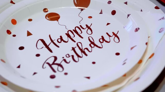 A stack of white paper plates with red "Happy Birthday" text and balloon designs sits on a white table indoors.