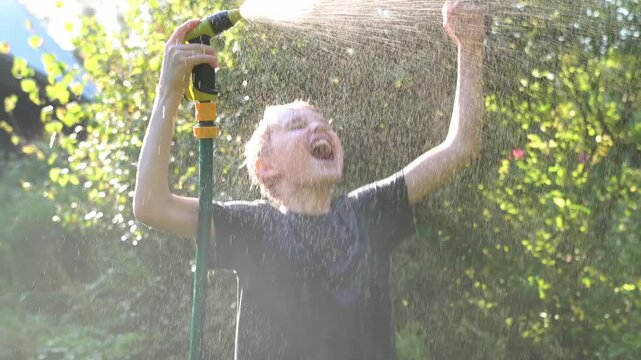 Funny boy pouring cool water on himself and playing with garden hose with sprinkler in sunny backyard on hot summer day. Happy child having fun with water splashes. Summer outdoor activity for kids.