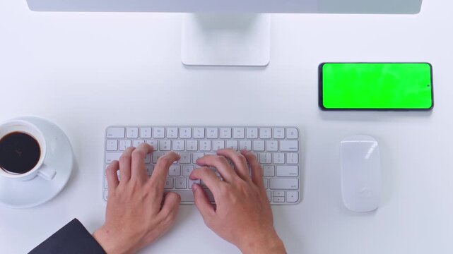 Top view of businessman typing on white keyboard at workdesk with green screen smartphone and coffee