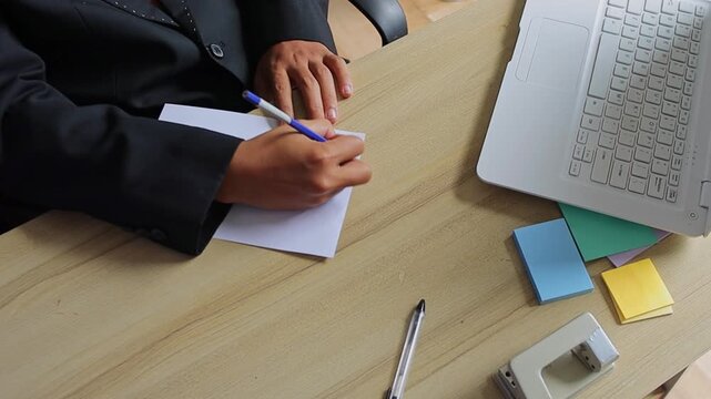 Top view of businessman in suit writing notes on white paper at wooden desk with office supplies