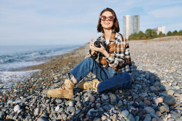 Fototapeta premium Photographer woman sits on a pebble beach with a camera, wearing a plaid shirt and boots, capturing a coastal scene under bright sun, conveying relaxed outdoor lifestyle and creative travel moments