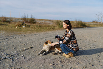 Naklejka premium Woman photographer kneels on a rocky desert path, focusing on a small dog while holding a camera, capturing candid moments in an open sunlit wide landscape.