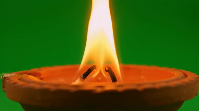 A close-up shot of a traditional clay lamp with twin wicks burning brightly, casting a warm, radiant flame against a vivid green screen background, symbolizing peace and illumination