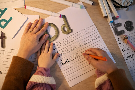Caucasian woman guiding Caucasian child writing letters on worksheet, adult hand helping child hand form letters, educational activity involving handwriting practice at desk