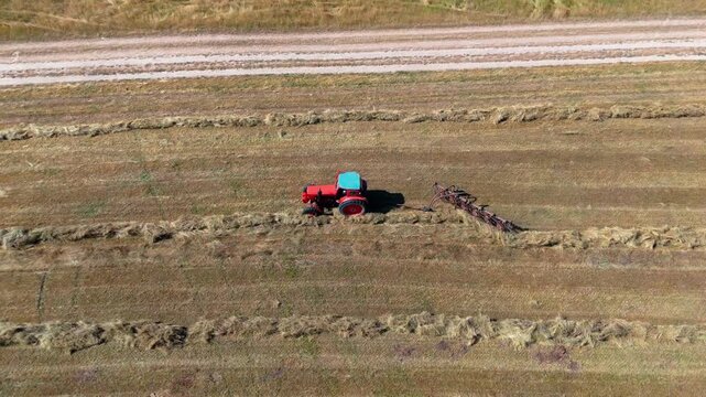 Top-down drone pan following a red tractor raking hay across cultivated farmland, emphasizing movement, symmetry, and agricultural patterns.