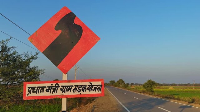 Tracking shot moving past a curve warning sign with Hindi text referencing Pradhan Mantri Gram Sadak Yojana, alongside a rural highway bordered by fields, trees, power lines, and clear blue sky.