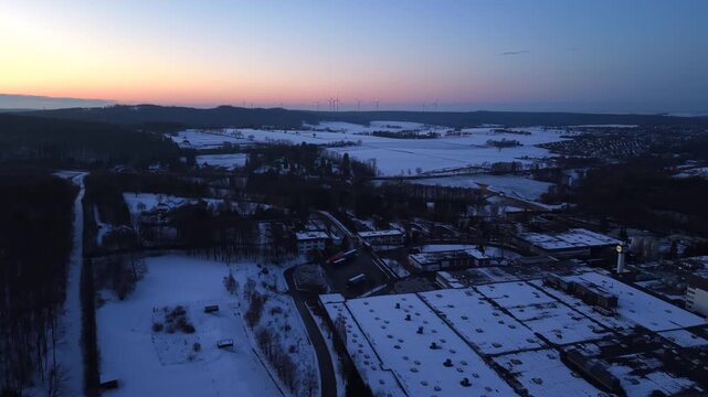 Aerial drone view of snow covered warehouse buildings in rural America at sunset, showing industrial rooftops, surrounding fields, distant hills and quiet winter atmosphere. Wide shot.