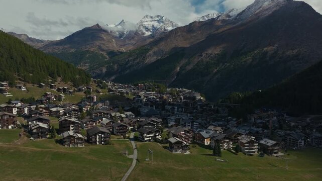 Aerial drone footage of the town of Saas-Fee with views over Allalin, Saas-Allmagell and Saas-Grund on a clear bluebird autumn day. Green valley floor contrasts with snow-covered alpine peaks.