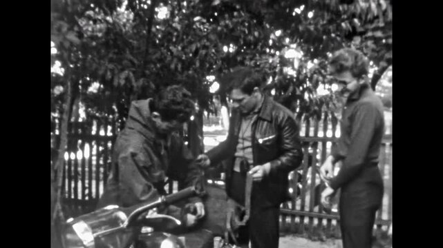 Men group preparing for summer motorcycle trip in backyard. Friends bikers attach travel bags to bike before departure. Adventure journey in motorbike. Old film. Archival retro 1970s. Vintage archive