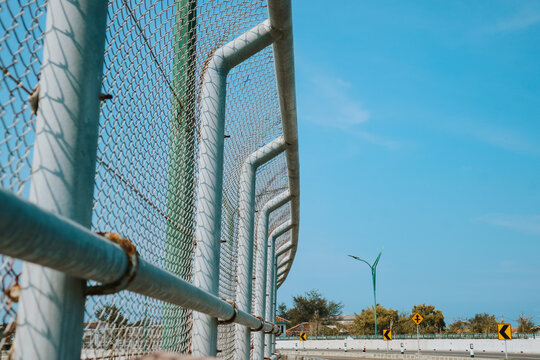 Close-up of curved chain-link fence supported by metal pipes along roadway, with clear blue sky, trees, and directional road signs in background, emphasizing repetitive pattern and perspective
