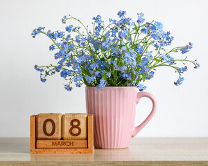 Wooden block calendar showing March 8 beside a pink ceramic mug filled with delicate blue forget me nots, celebrating International Women's Day and springtime