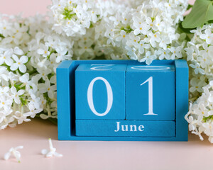 Wooden calendar block showing the first day of June, surrounded by fresh white lilac flowers on a light pink surface, symbolizing the beginning of summer