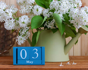 White lilac branches with green leaves blooming in a light green watering can, sitting on a wooden table next to a blue block calendar showing May 3