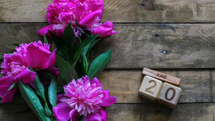 Fototapeta premium Pink peony flowers with green leaves decorating a rustic wooden background, featuring wooden block calendar displaying the date June 20, symbolizing summer and special occasions