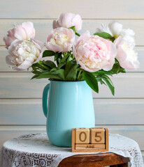 Pink peony flowers in a blue pitcher sit on a wooden stool with a lace tablecloth next to a wooden block calendar showing June 5th, creating a serene floral still life