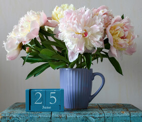 Peonies in a purple ribbed mug beside a blue wooden calendar block showing June 25 on a weathered turquoise table, a soft still life evoking summer and celebration