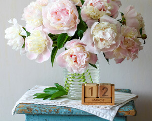 Pink peonies blooming in a glass vase placed on a vintage stool with a wooden block calendar displaying March 12, symbolizing the arrival of spring and fresh beginnings