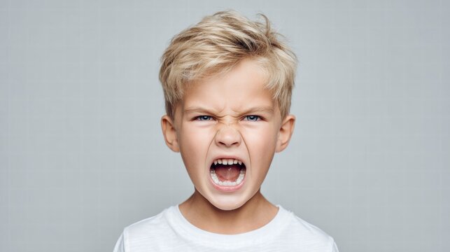 Angry young boy with blonde hair yelling with mouth wide open against a gray background.