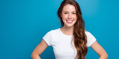 Obraz premium Smiling woman with long brown hair wearing white t-shirt poses confidently against blue background in bright and cheerful atmosphere for lifestyle concepts