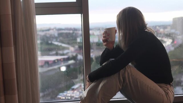 Young woman sitting on a windowsill and drinking coffee while looking at the city through a rainy window. Cozy home lifestyle scene with soft natural light, calm mood, urban view and quiet moment of