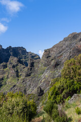 Rocky mountain gorge near Curral das Freiras Madeira, rugged volcanic cliffs with green heath shrubs and distant peaks beneath clear blue sky and drifting cloud