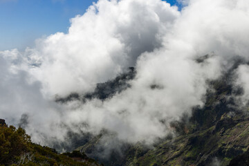 Fototapeta premium Rugged cliffs and green shrub slopes above Faial Santana in Madeira, rolling cumulus clouds spill across the ridge line and hide distant peaks in bright daylight