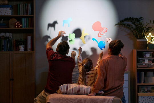 Caucasian man, Caucasian woman, and young Caucasian girl sitting together creating animal shadow puppets on wall using colorful cutouts, engaging in playful family activity at home