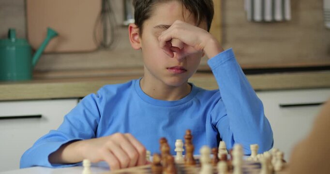 Smart boy thinks about next move in chess game. Concentrated child plays chess with father at home. Kid learns strategy and logic. Intellectual board game in kitchen. Family game of chess.