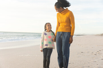 Happy mother and daughter holding hands while looking at each other on beach against sky