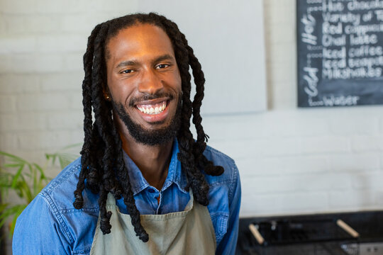 African American man smiling in coffee shop wearing denim shirt and grey apron with chalkboard menu