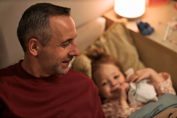Obraz premium Middle aged Caucasian man sitting on bed smiling while young Caucasian child lying next to him holding blanket, both relaxing together in cozy bedroom setting during evening