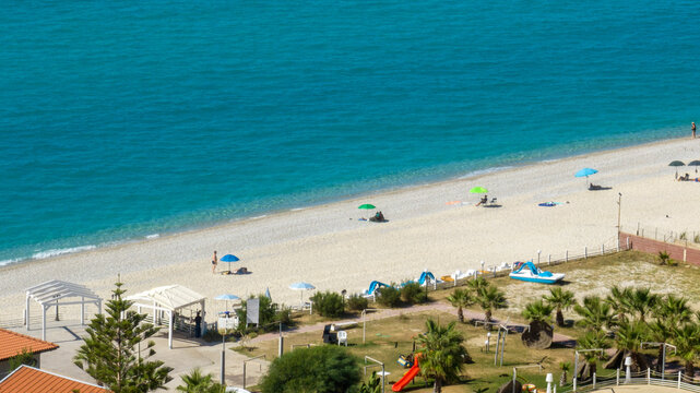 Aerial view of a wide sandy beach meeting a bright turquoise sea. Scattered colorful umbrellas and small white structures dot the shoreline. It is Zambrone beach, near Tropea, in Calabria, Italy.