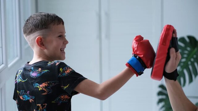 A child wearing boxing gloves practices boxing with his father. The boy punches the boxing mitt.