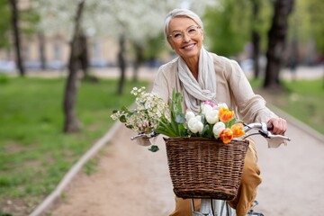 Fototapeta premium Cheerful senior woman riding a vintage bicycle with a flower basket in a spring park representing active aging and carefree retirement